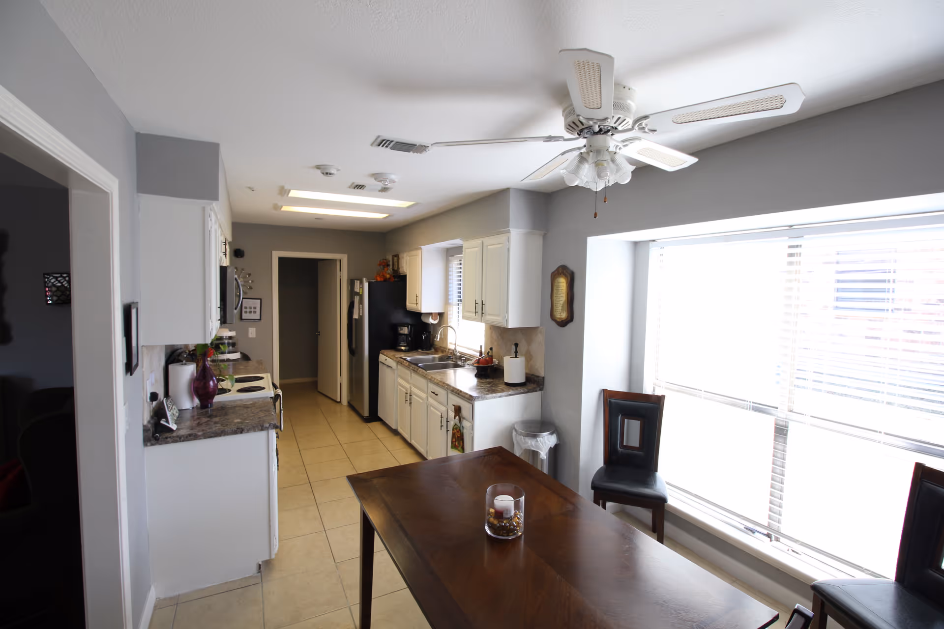 Bright galley kitchen with white cabinets, countertop appliances, a ceiling fan, and a wooden dining table by a large window.