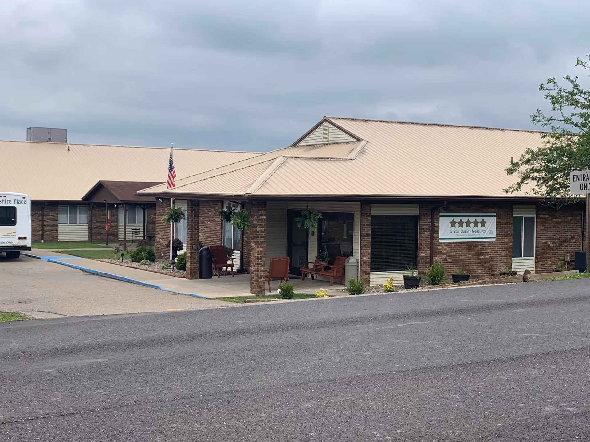 Exterior view of a single-story brick building with a tan metal roof, a covered porch with seating, hanging plants, and an American flag. A white bus labeled 'Abbyshire Place' is parked on the left side. There is a sign on the building displaying five stars and the text '5 Star Quality Measures'. The sky is overcast.