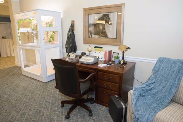A cozy sitting area with a wooden desk and chair, vintage telephone and typewriter, framed horse artwork, and a nearby couch with a blue throw.