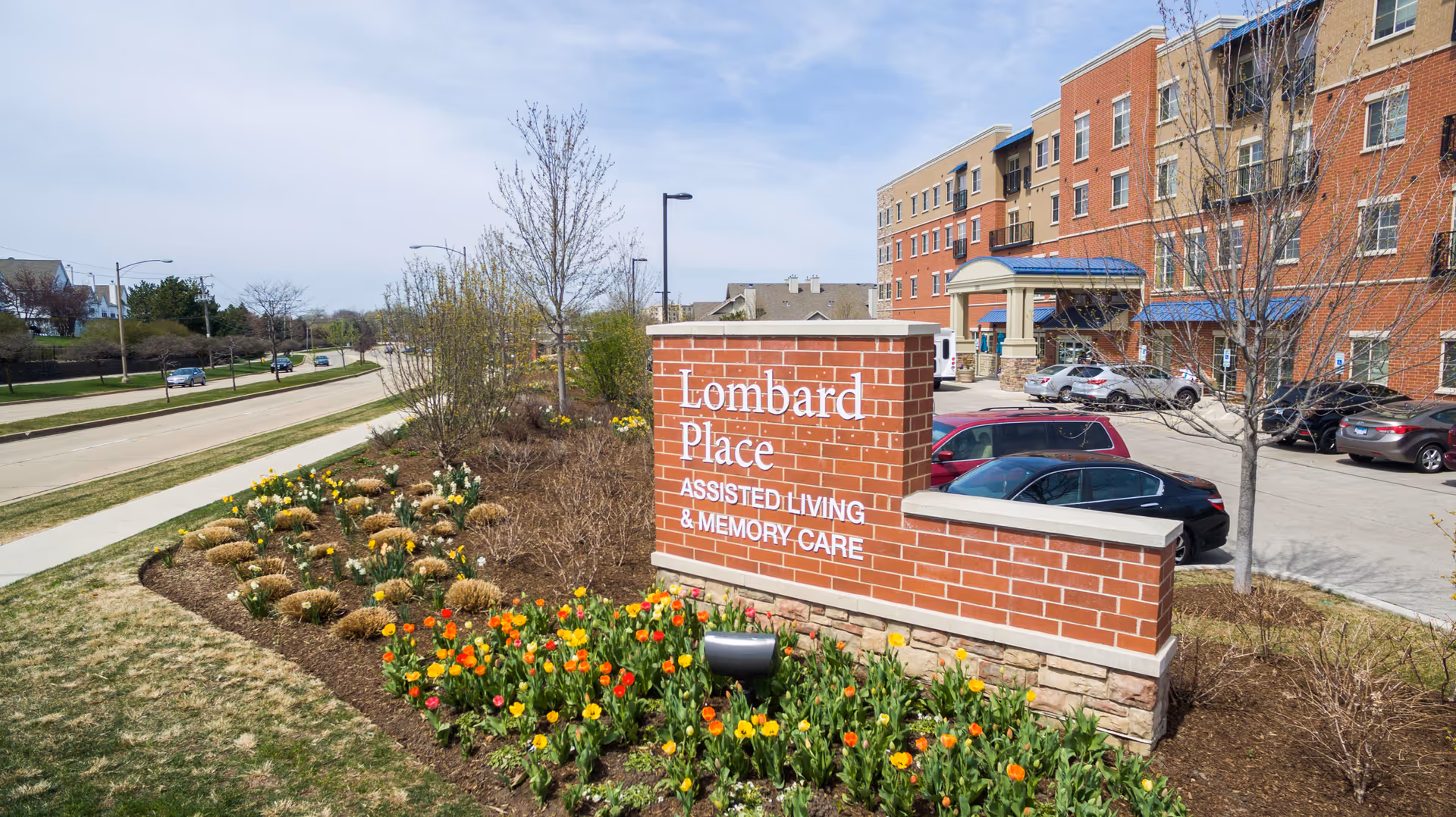 Outdoor view of Lombard Place Assisted Living & Memory Care sign made of brick with flowers planted in front. A multi-story brick building and parked cars are visible in the background along a street with sidewalks and trees.