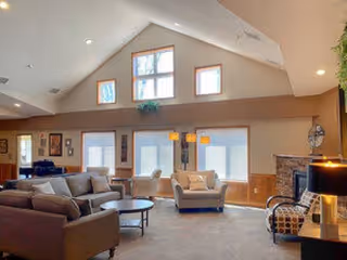 A spacious living room with high vaulted ceilings and multiple windows letting in natural light. The room features a large sectional sofa, two armchairs, a coffee table, a fireplace with a stone surround, and various lamps and decorative items. The walls are painted in neutral tones with wooden trim around the windows.