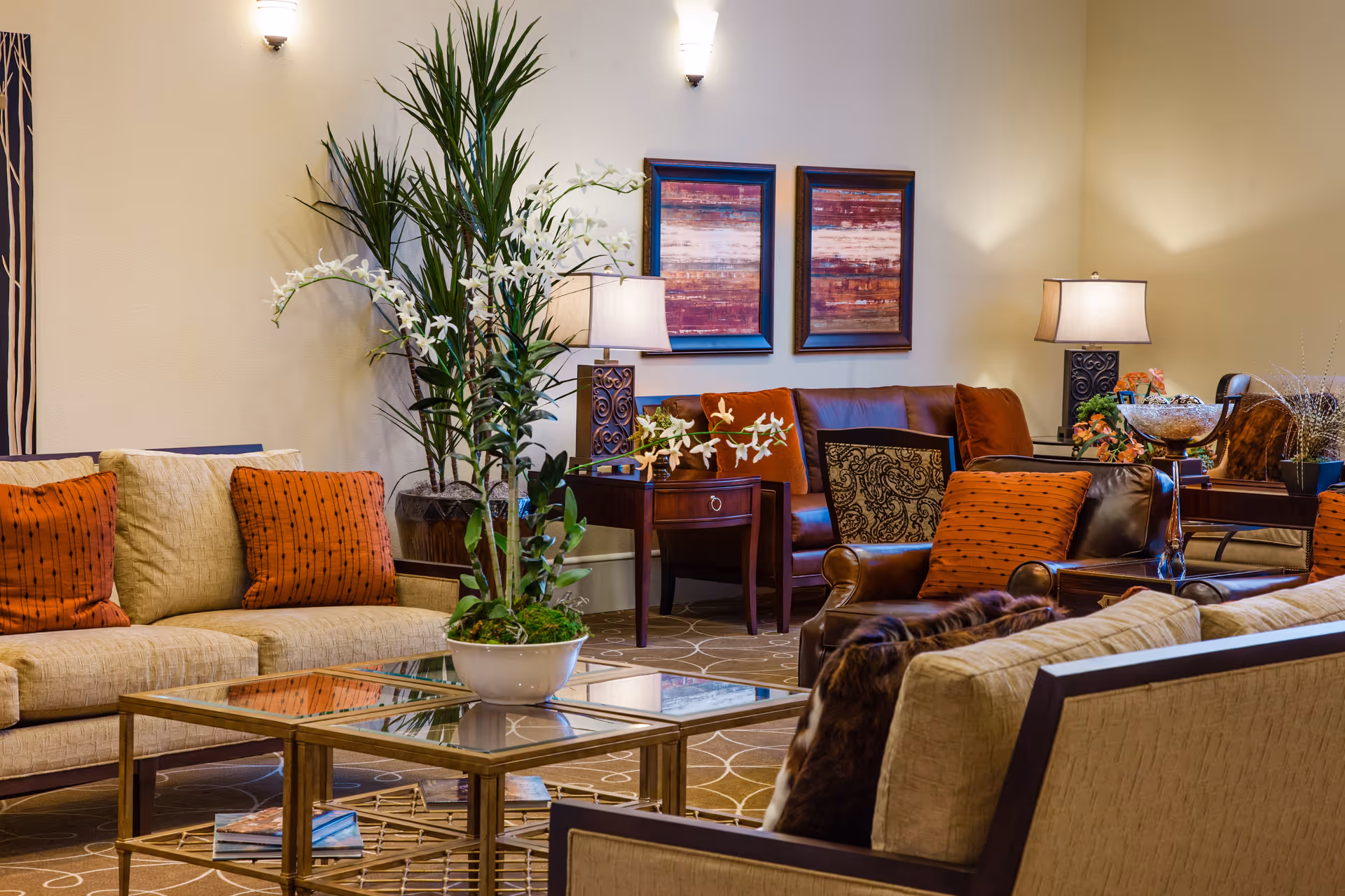 A cozy living room area with beige and brown sofas adorned with orange and patterned cushions. There is a glass coffee table with a potted plant featuring white flowers in the center. The room is decorated with two framed abstract paintings on the wall, two table lamps, and a tall green plant in the corner.