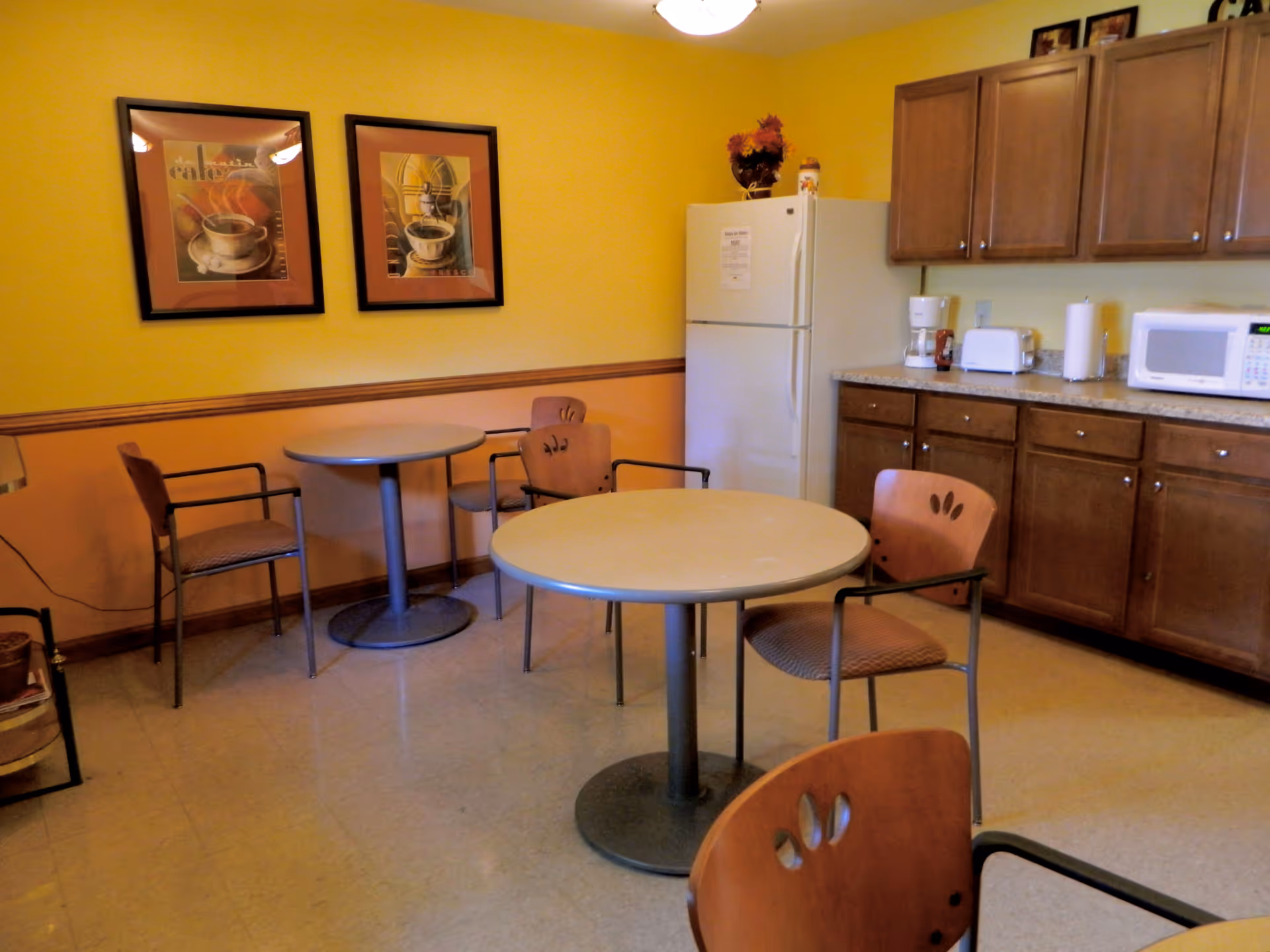 Small communal dining area with round tables and chairs, yellow walls, and a kitchenette with a refrigerator, microwave, and cabinets.