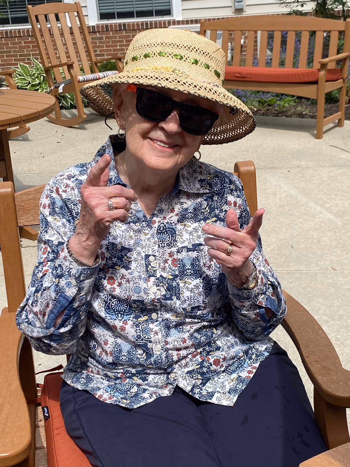 An elderly woman wearing a patterned shirt, dark sunglasses, and a straw hat is sitting outdoors on a wooden chair with an orange cushion. She is smiling and making finger gun gestures with both hands. Behind her, there are more wooden chairs and a bench with an orange cushion, along with some plants and a brick wall.