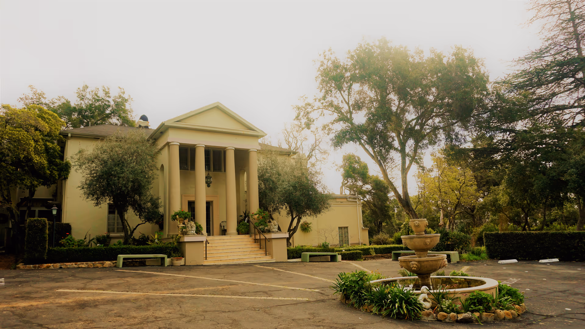 Front view of a beige senior living facility with columns, steps, and a fountain.
