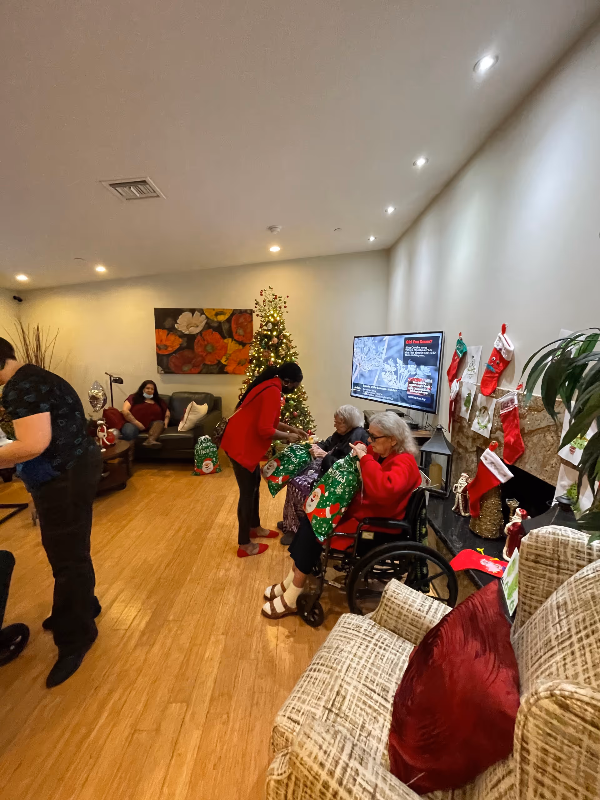 Residents and staff exchanging holiday gifts in a decorated common room with a Christmas tree, stockings, and seating.