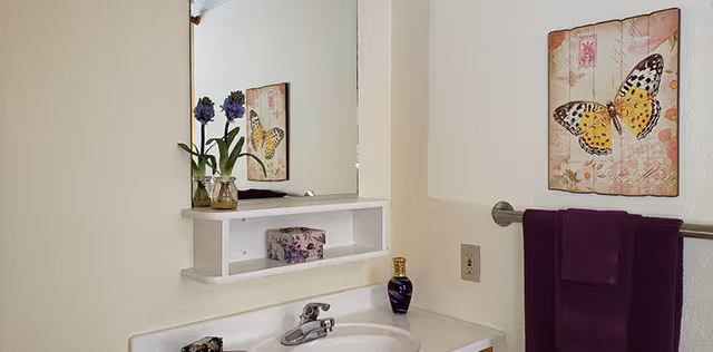 Bathroom sink and mirror with a small shelf holding decorative flowers and a box, butterfly wall art, and a purple towel on a towel bar.