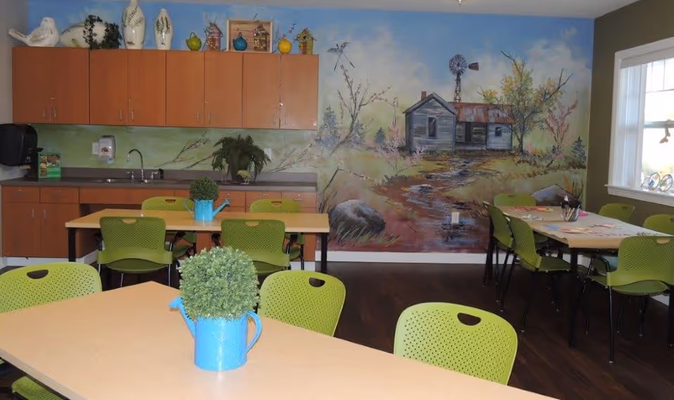A bright room with light wooden tables and green chairs arranged for group seating. The back wall features a mural of a rustic house with trees and a windmill. Above a countertop with a sink are wooden cabinets decorated with various vases and birdhouses. Small potted plants in blue watering can containers are placed on the tables. A window on the right side lets in natural light.