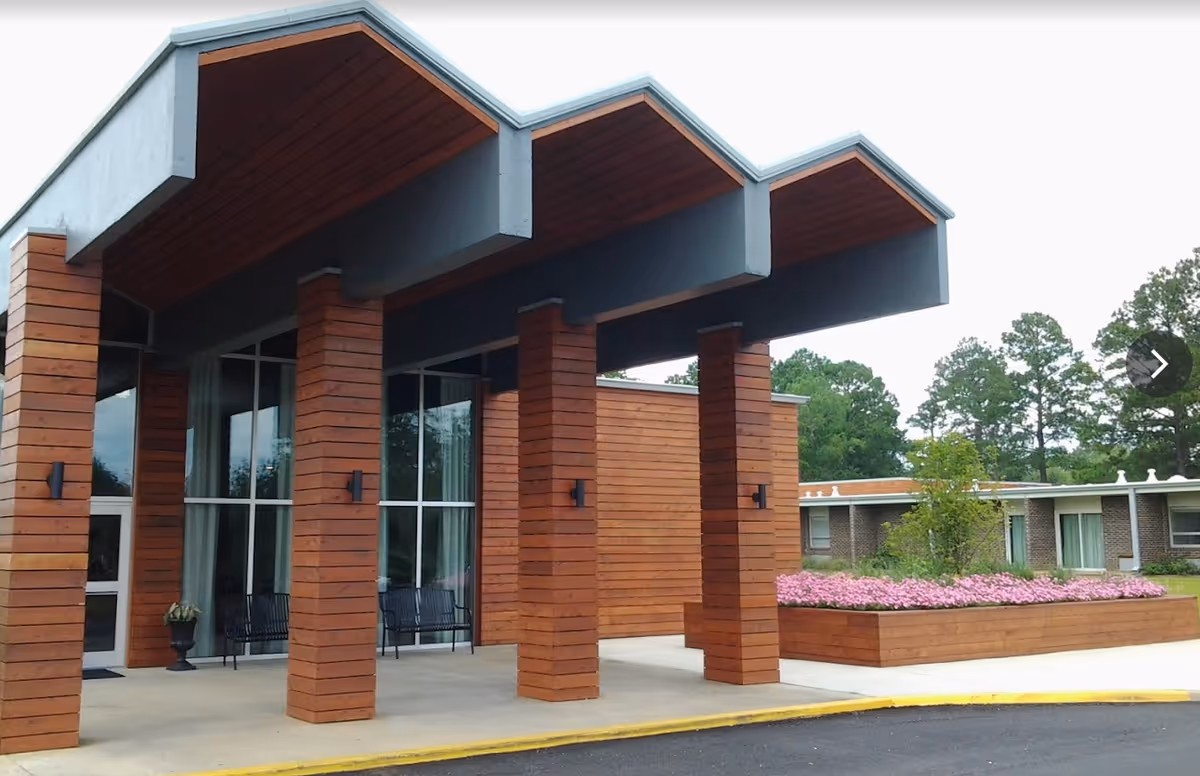 Entrance area of Wesley Place on Honeysuckle featuring a modern wooden canopy supported by four wooden pillars, with large glass windows and a flower bed filled with pink flowers to the right.