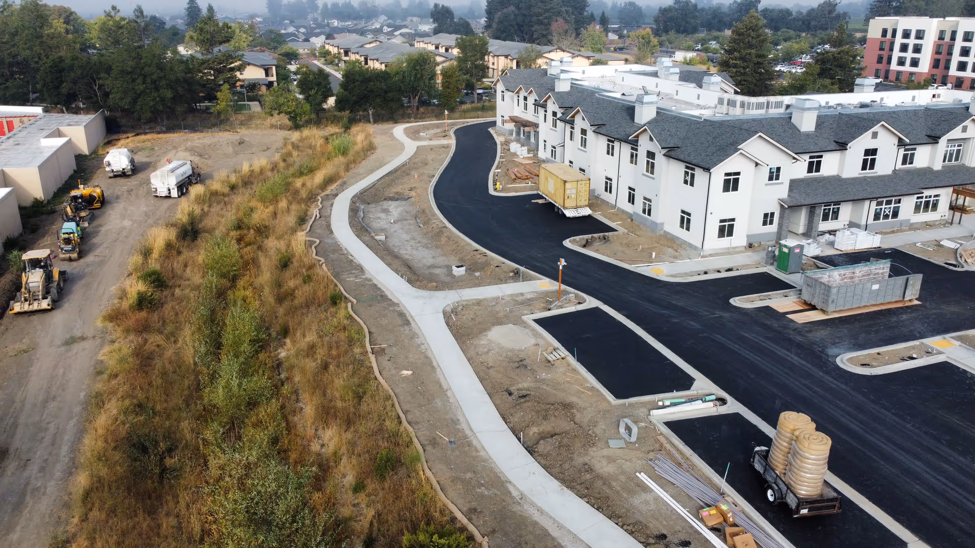 Aerial view of a senior living facility under construction with a newly paved road and sidewalks. The building is a multi-story structure with white exterior walls and dark roofing. Construction materials and equipment are visible around the site, along with some landscaping and grassy areas on the left side.