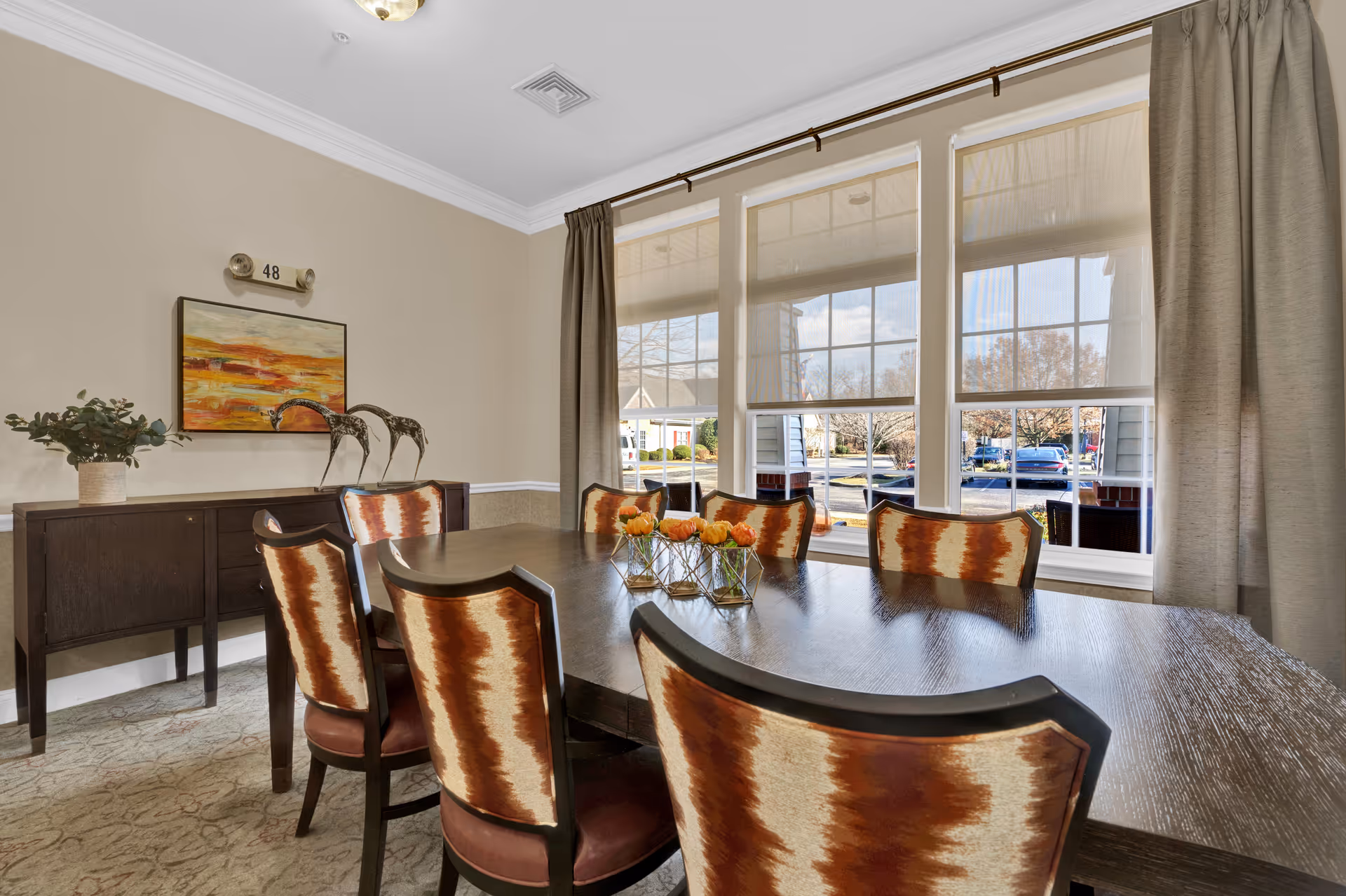 A dining room with a dark wooden table surrounded by eight chairs with patterned upholstery in shades of brown and cream. On the table are three small glass vases with orange flowers. Behind the table is a dark wooden sideboard with two decorative giraffe sculptures and a potted plant. Above the sideboard hangs a colorful abstract painting. Large windows with beige curtains let in natural light and show a view of parked cars and trees outside.