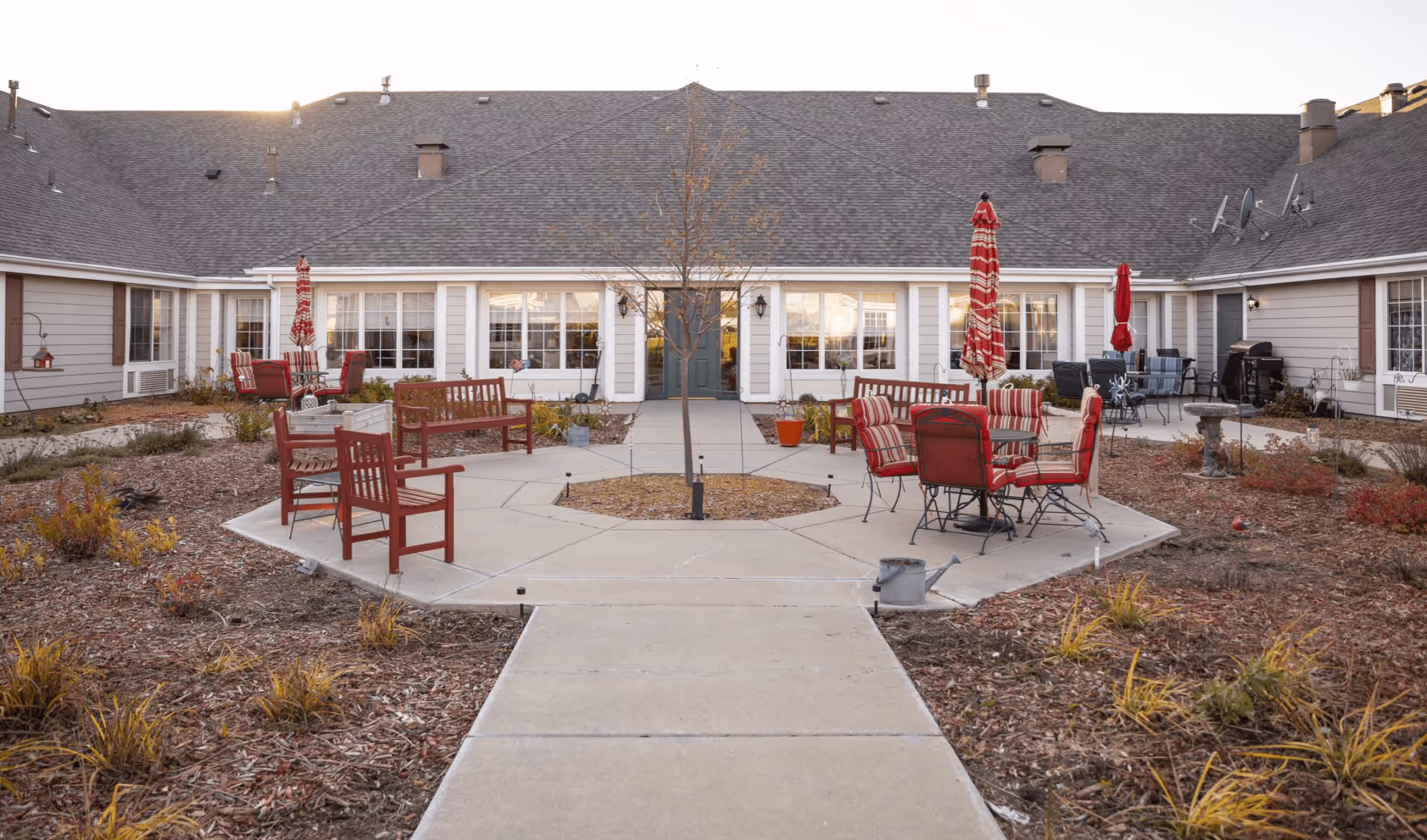 Outdoor courtyard area of a senior living facility with a central paved walkway leading to a small tree surrounded by benches and patio tables with red chairs and umbrellas. The building surrounds the courtyard with large windows and a central door.