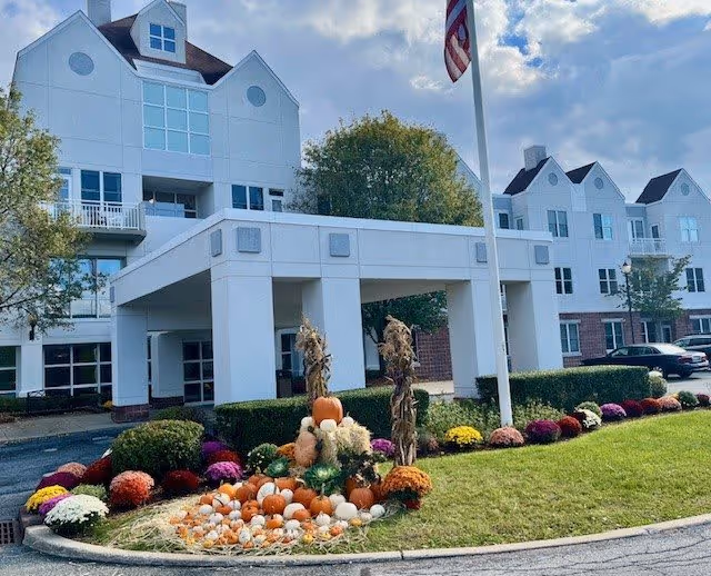 Exterior view of a multi-story senior living facility with white walls and multiple windows. The entrance has a covered drop-off area with columns. In front of the entrance, there is a landscaped area decorated with pumpkins, gourds, hay, and colorful flowers, along with two dried cornstalks. An American flag is flying on a flagpole near the entrance. Trees and parked cars are visible in the background under a partly cloudy sky.