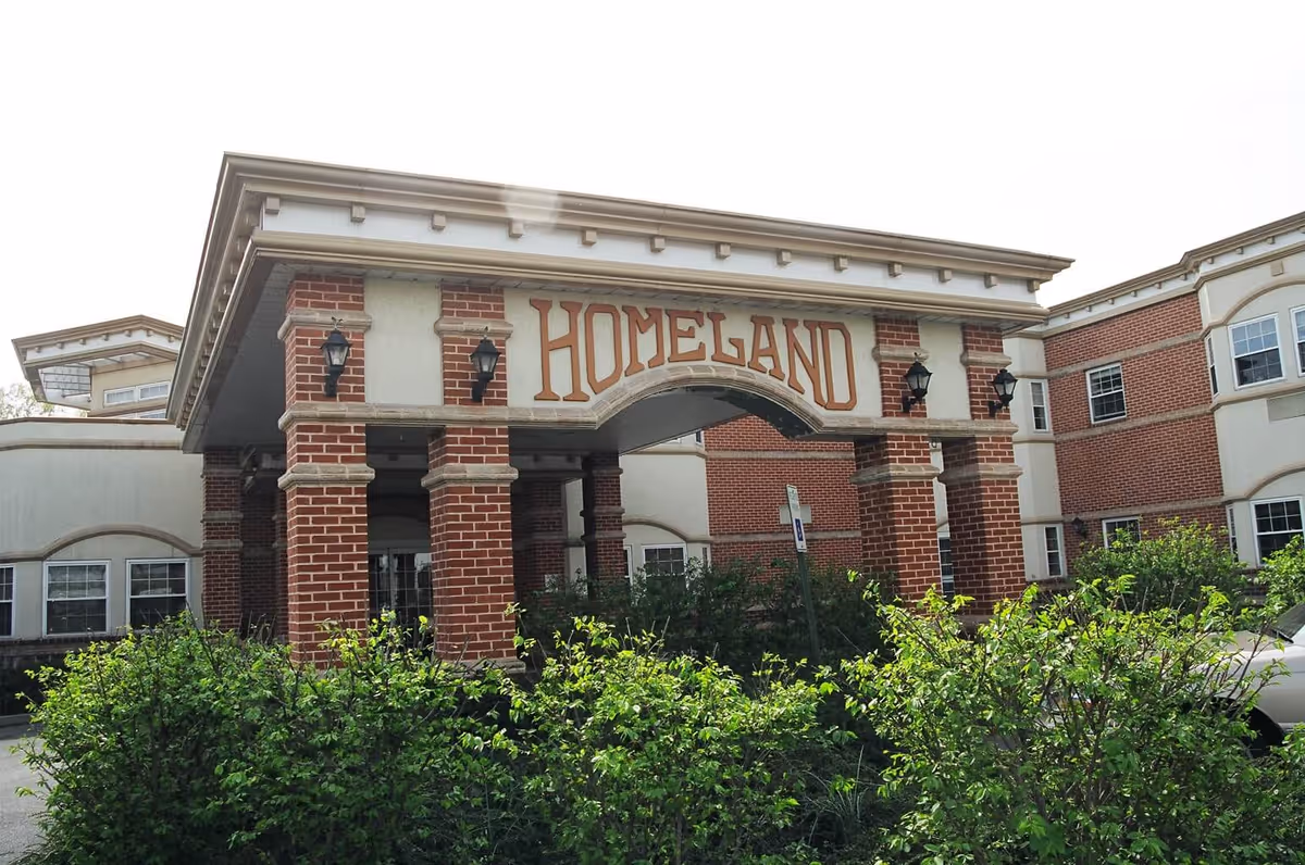Front entrance of a brick senior living building with a portico sign reading "HOMELAND" and shrubs in the foreground.