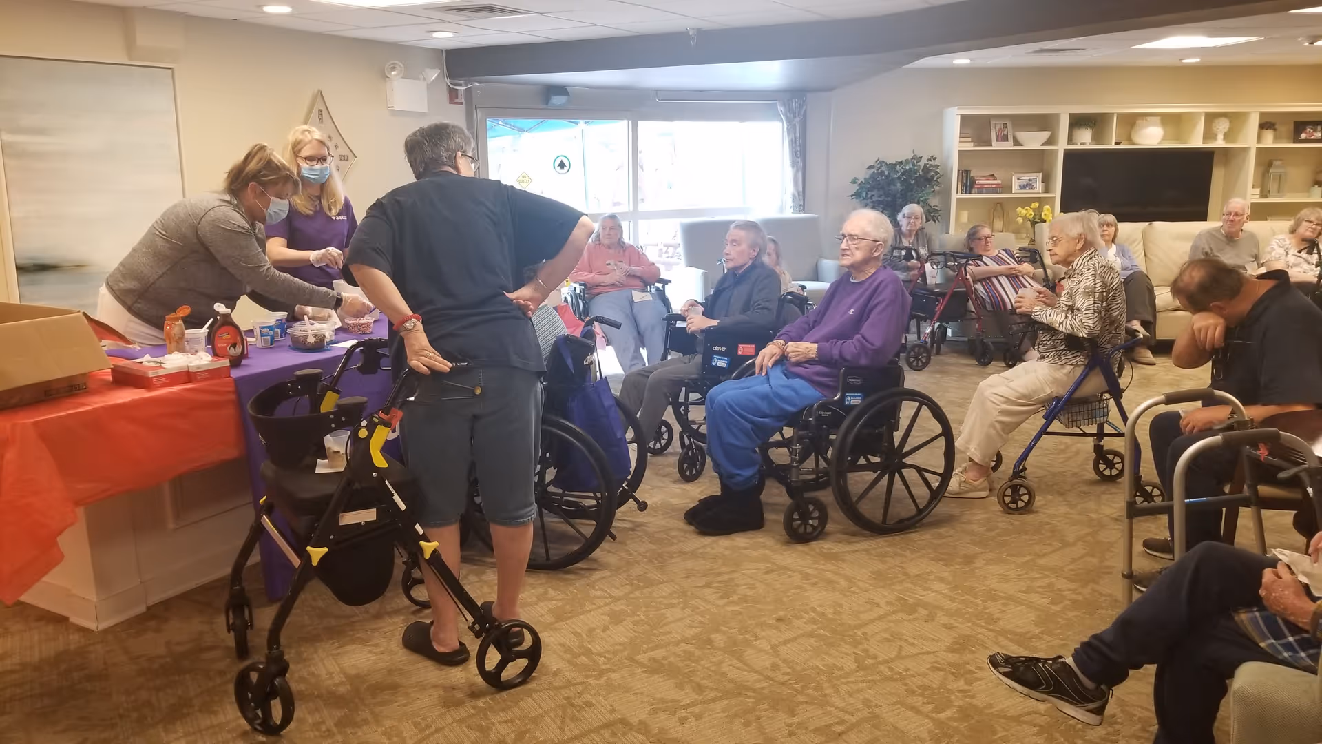 A group of elderly people, some in wheelchairs and others with walkers, gathered in a common room. Two caregivers wearing masks are serving food or snacks at a table covered with a red and purple tablecloth. The room has beige carpet, shelves with decorations, and large windows letting in natural light.