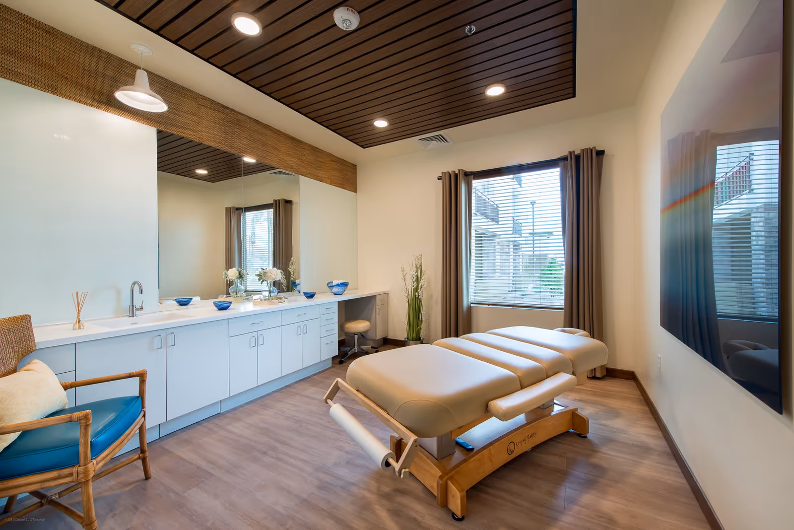 A bright and clean therapy or massage room with a padded treatment table in the center, a large mirror on the wall above a long white cabinet with a sink, decorative bowls, and flowers. There is a comfortable chair with a cushion, a window with brown curtains, and a wooden ceiling panel with recessed lighting.