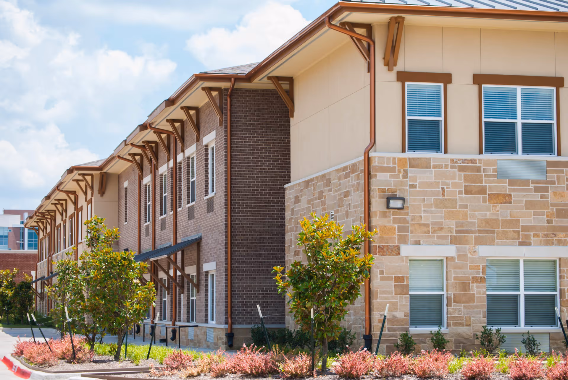 Exterior view of a two-story assisted living facility building with a combination of brick and stone facade, multiple windows, small trees, and landscaped bushes along the sidewalk under a partly cloudy sky.