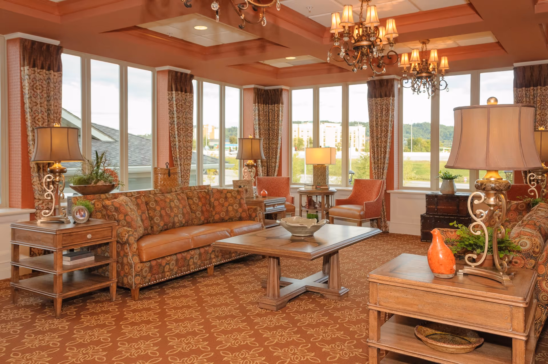Sunlit, upscale communal living room with patterned sofas and armchairs around a wooden coffee table under chandeliers.