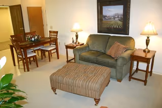 A living room area with a green upholstered sofa, a striped ottoman, two wooden side tables each with a lamp, and a wooden dining table with four chairs. A framed landscape painting hangs on the wall above the sofa.
