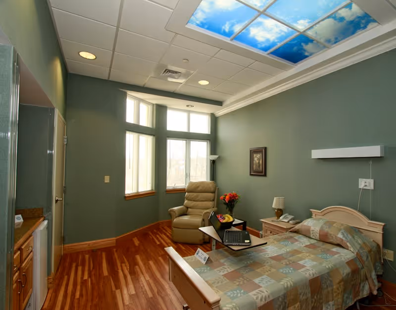 A furnished senior bedroom with a single bed, bedside table, reclining chair, windows, and a laptop on a tray under a ceiling sky panel.