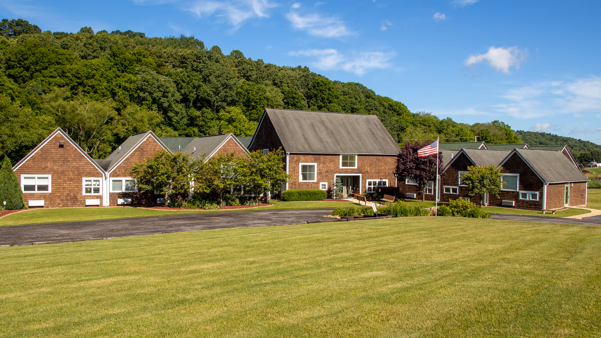 Exterior view of a single-story personal care home building with brown wooden siding and multiple gabled roofs, surrounded by a well-maintained green lawn and trees, with a clear blue sky in the background.