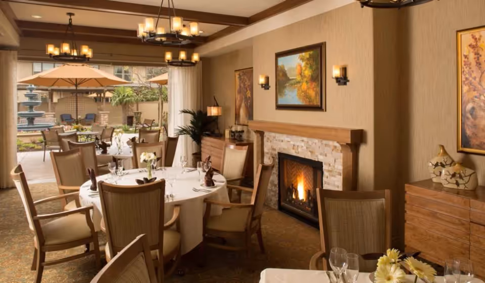 A cozy dining room with round tables covered in white tablecloths, set with glasses, napkins, and flowers. The room features a lit fireplace with a stone surround, wooden furniture, wall sconces, and framed paintings. Large windows and doors open to an outdoor patio area with tables, chairs, umbrellas, and a water fountain.