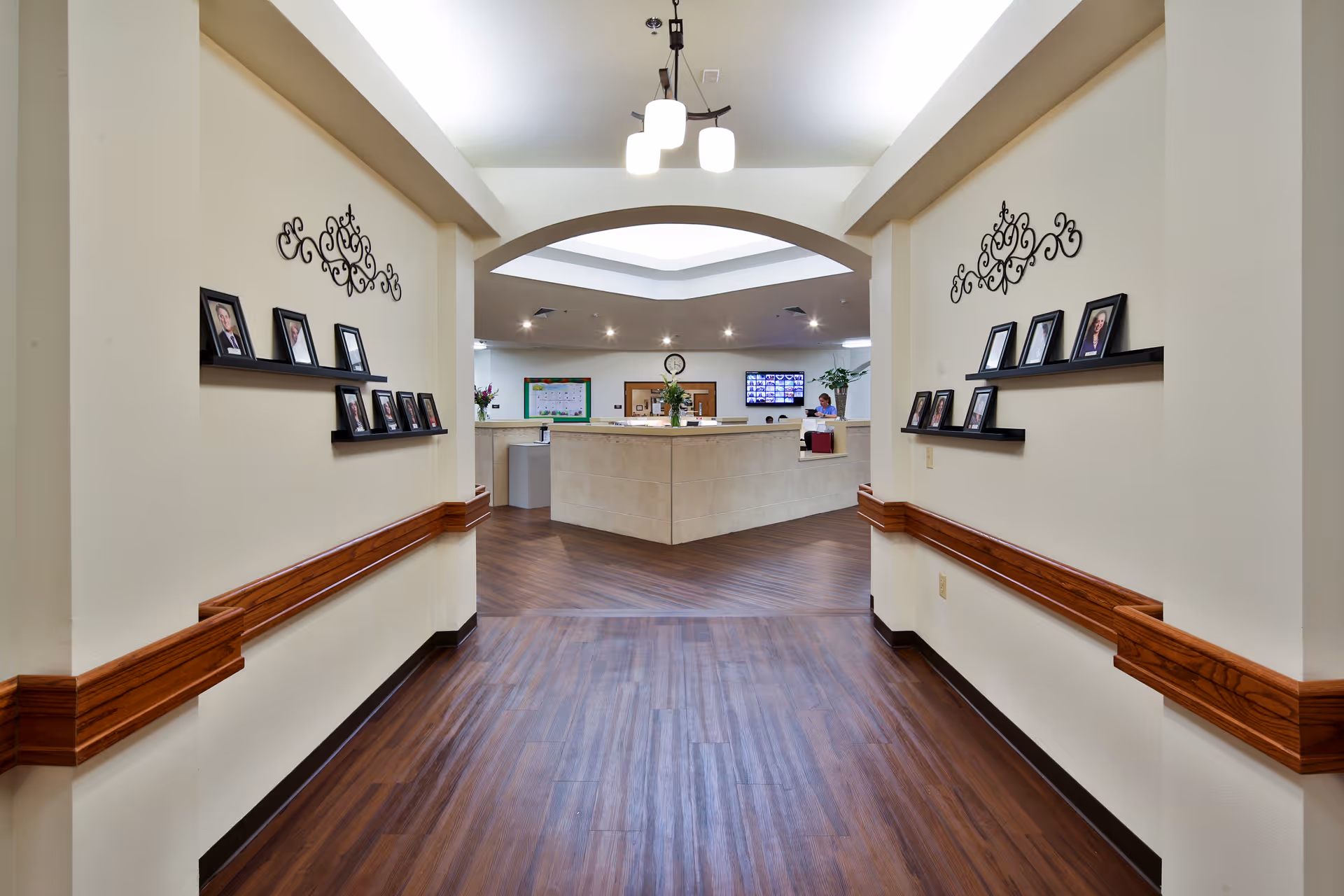 Interior hallway of a senior living facility with wood flooring and cream-colored walls. The hallway leads to a reception desk area under a skylight. Both walls of the hallway have small black shelves displaying framed photos and decorative metal wall art above them. A ceiling light fixture hangs in the hallway.