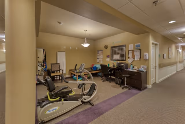 Interior view of a senior living facility hallway with a small exercise area on the left, featuring a NuStep exercise machine, chairs, a desk with office supplies, and a bulletin board on the wall. The hallway extends to the right with doors and handrails along the wall.