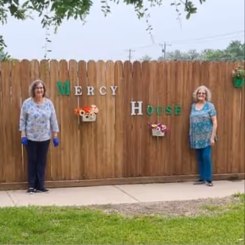 Two women stand on either side of a wooden fence decorated with letters and small planters spelling "Mercy House".