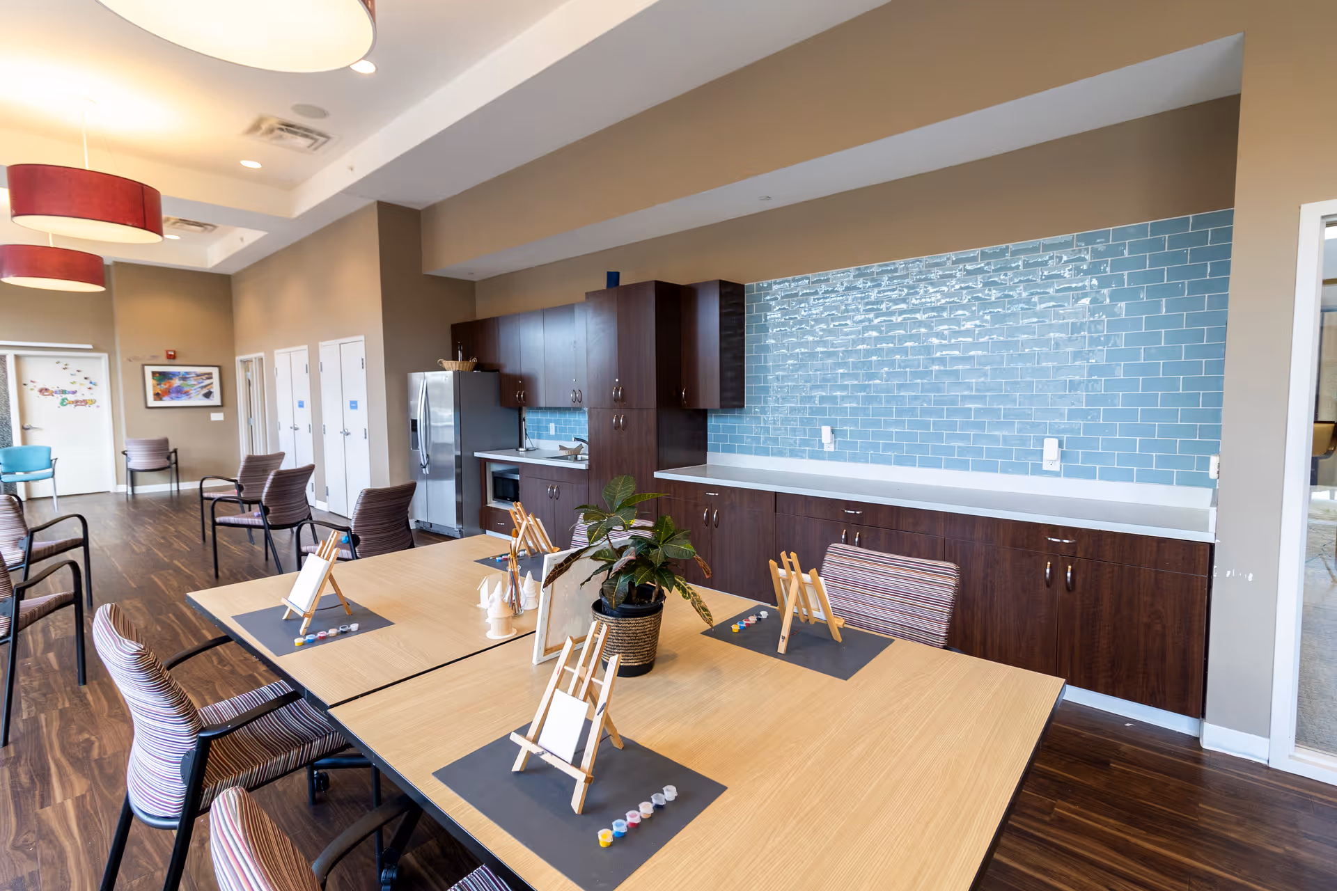 A spacious activity room with wooden tables and striped chairs arranged around them. Each table has small easels with blank canvases and paint sets, suggesting an art activity setup. The room features a kitchenette area with dark wood cabinets, a stainless steel refrigerator, a microwave, and a blue tiled backsplash. The floor is wooden, and there are large hanging light fixtures with red lampshades. Additional chairs are placed along the wall, and there is a door with colorful decorations in the background.