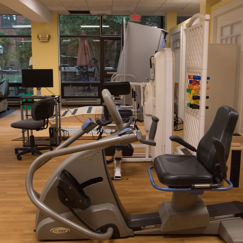 Indoor exercise room with various fitness equipment including a seated recumbent bike, weight machine, and a computer workstation. Large windows show an outdoor patio area with trees and umbrellas.