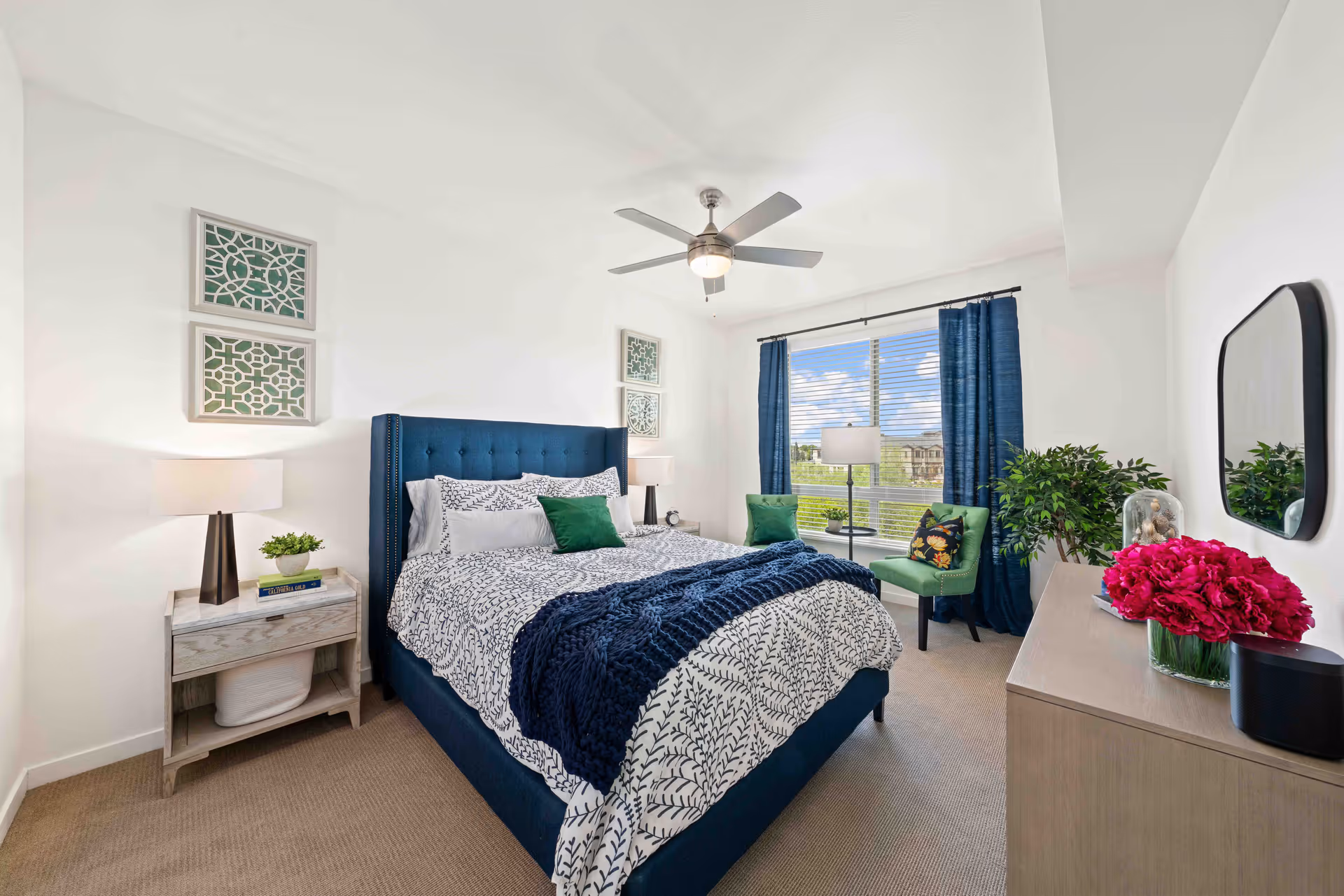 A bright and neatly arranged bedroom featuring a large bed with a blue upholstered headboard, white and blue patterned bedding, and green accent pillows. The room has a ceiling fan with light, two framed decorative wall art pieces above the bed, a nightstand with a lamp and small plant, a window with blue curtains letting in natural light, a green chair with a floral cushion, a floor lamp, a dresser with a mirror, and a vase of pink flowers.