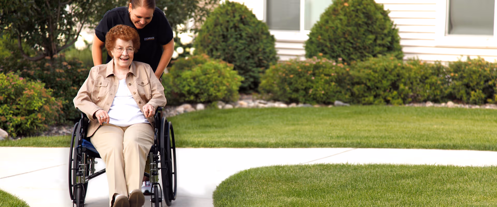 An elderly woman in a wheelchair is being pushed by a caregiver along a paved pathway outside. They are both smiling and surrounded by green grass, bushes, and trees with a building visible in the background.
