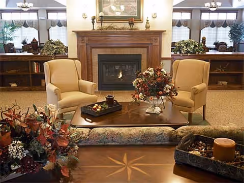 A cozy living room area featuring two beige armchairs facing a wooden coffee table with floral arrangements and decorative items. Behind the chairs is a fireplace with a wooden mantel, adorned with candles and a framed painting above it. The room has large windows with curtains and plants on either side, creating a warm and inviting atmosphere.