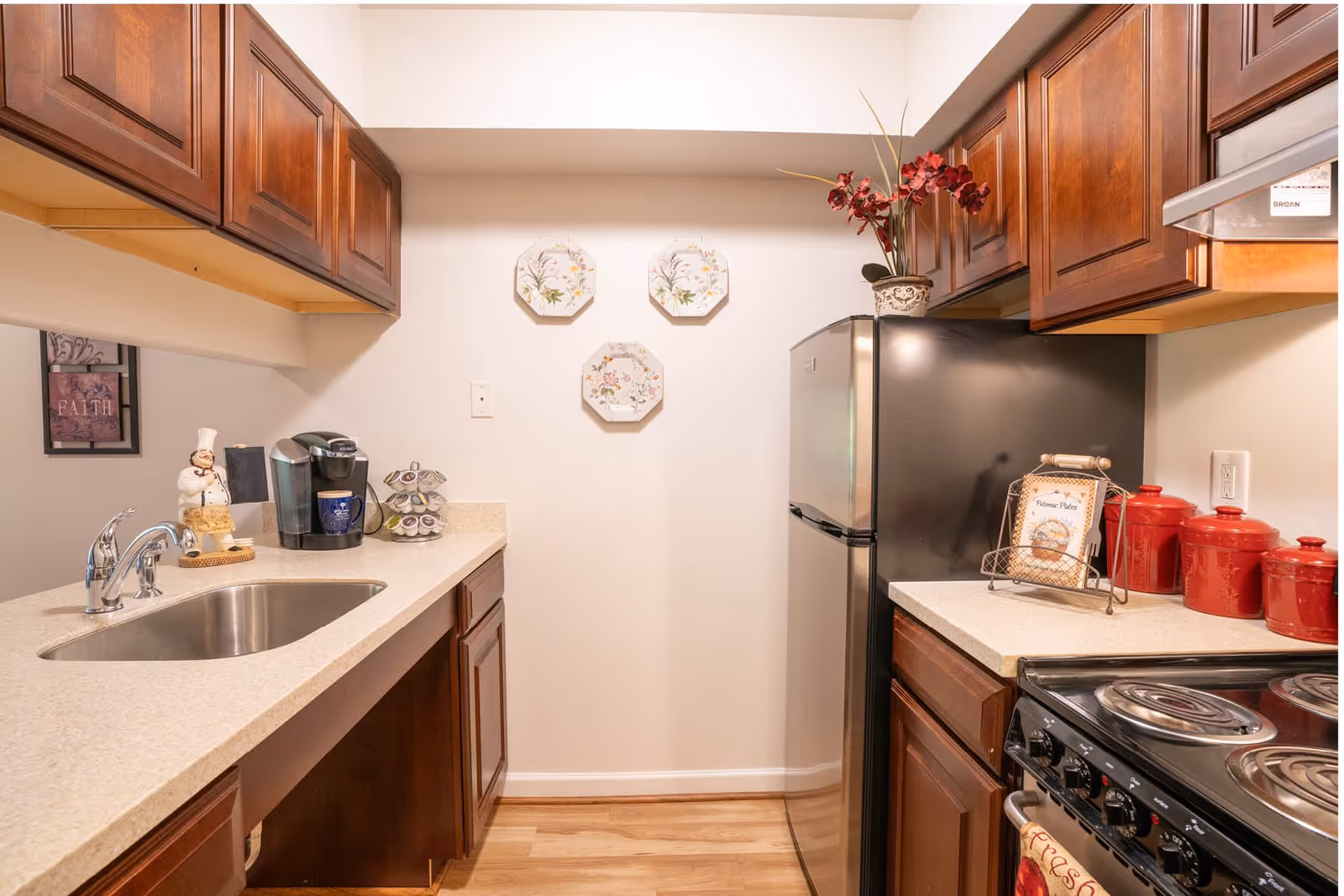A compact kitchen with wooden cabinets, a stainless steel sink, a black refrigerator, and an electric stove with coil burners. The countertop holds a coffee maker, a decorative chef figurine, a spice rack, red canisters, and a small framed sign. Three decorative floral plates hang on the wall above the refrigerator, and a vase with red flowers sits on top of the fridge.