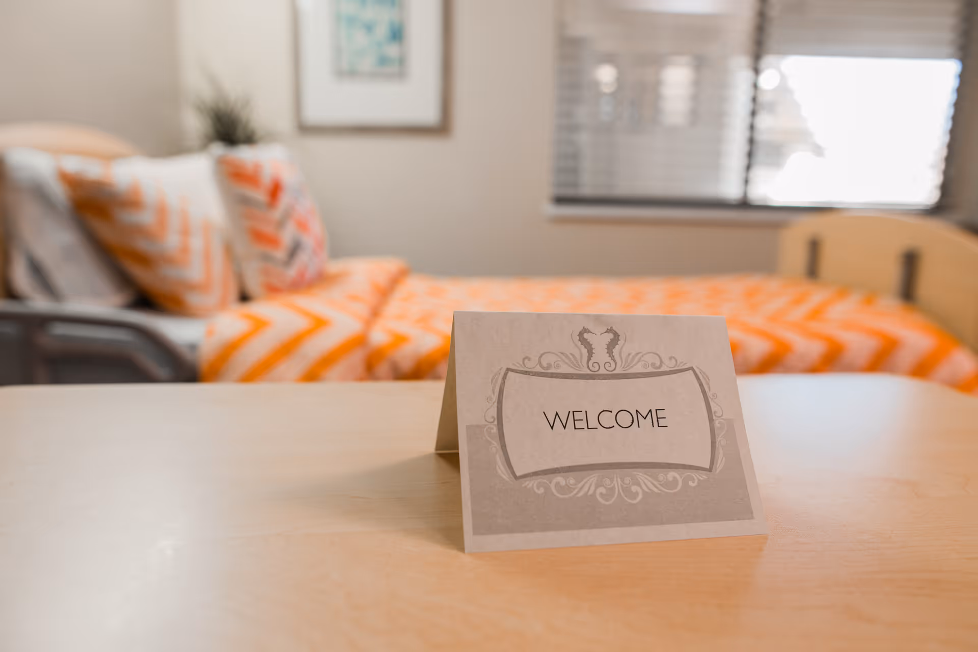 A welcome tent card on a table in front of a bed with orange chevron bedding and a window in a bright room.