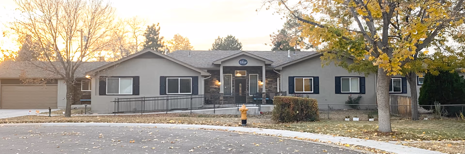 Front exterior view of a single-story senior living facility building with a gray facade, black window shutters, and a central entrance with a small porch. There are trees with autumn leaves and a fire hydrant in front of the building, along with a curved driveway.