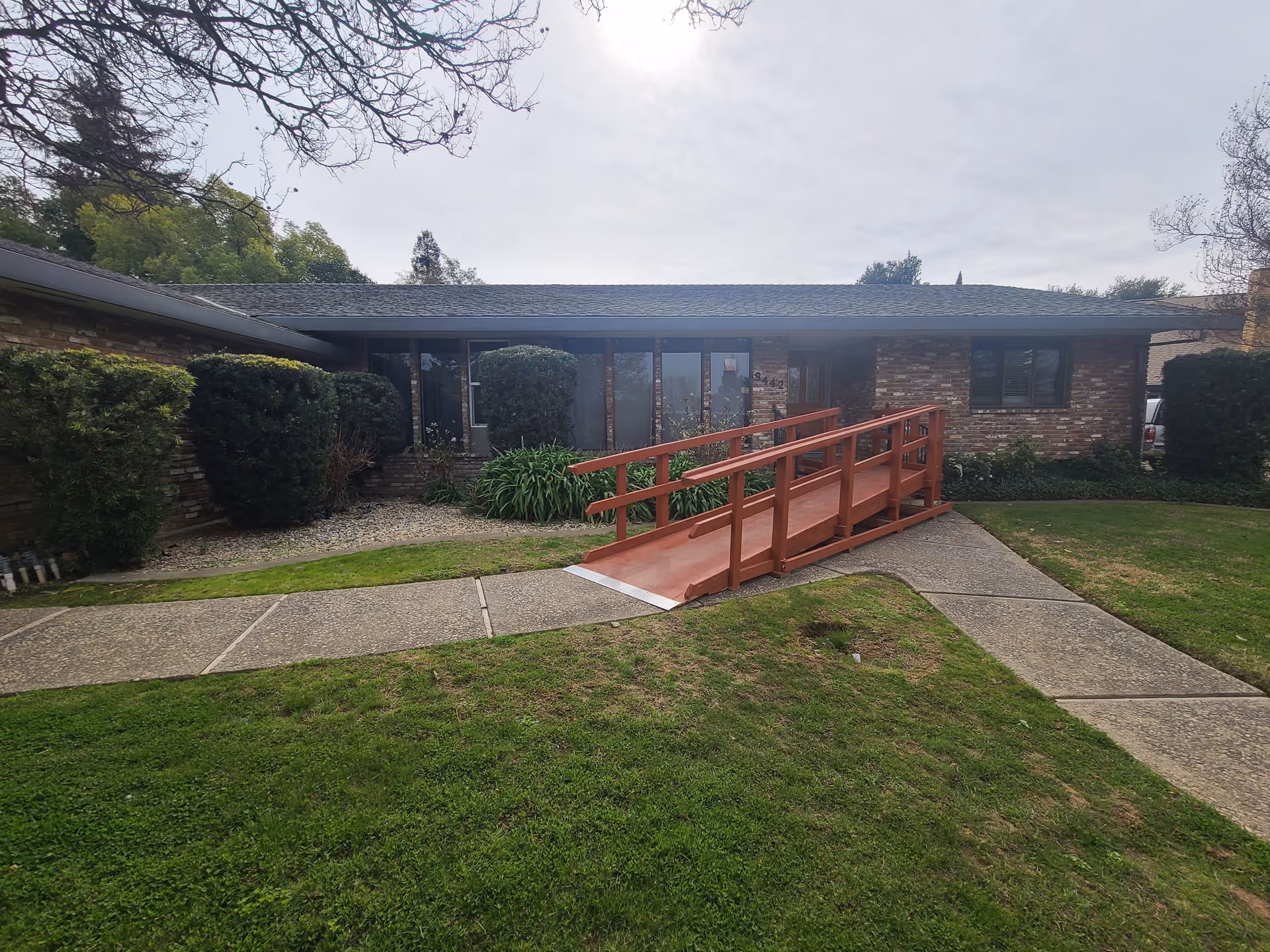 Front exterior of a single-story brick building with a red wooden wheelchair ramp leading to the entrance and a lawn with shrubs.