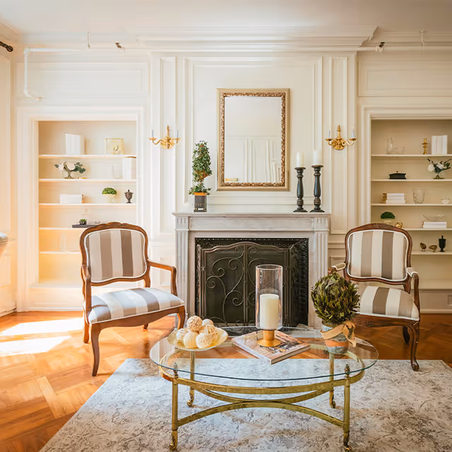A bright and elegant living room with two striped armchairs facing a glass coffee table. The table holds decorative items including a candle, a small plant, and a plate with decorative balls. Behind the chairs is a fireplace with a decorative screen, topped with a mirror, two black candle holders with white candles, and a small potted plant. Built-in shelves on either side of the fireplace hold various decorative objects and books. The room features white paneled walls, wooden parquet flooring, and a light-colored area rug.