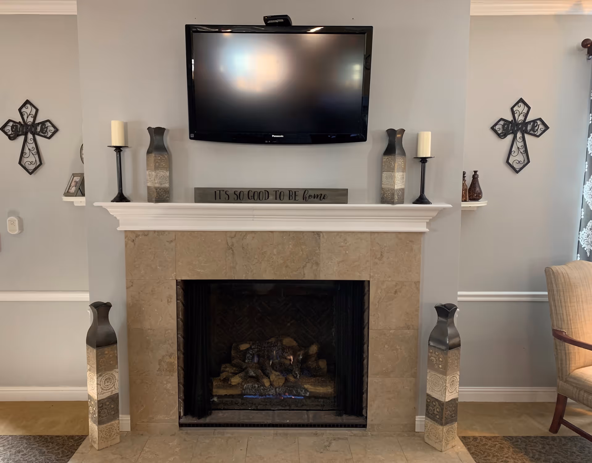 A cozy living room area featuring a beige tiled fireplace with a mounted flat-screen TV above it. On the mantel, there are two decorative candle holders with white candles and two tall decorative vases. A wooden sign on the mantel reads 'IT'S SO GOOD TO BE home'. On either side of the fireplace, there are decorative crosses mounted on the light gray walls. A beige upholstered chair is partially visible on the right side of the image.