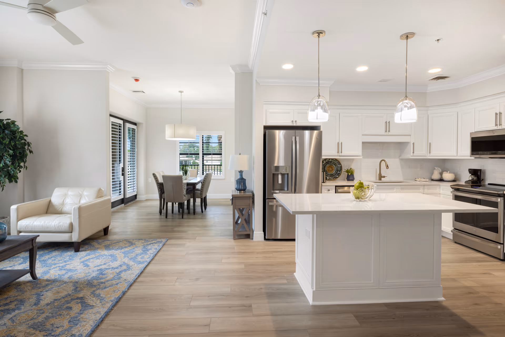 Bright and modern open-concept living space featuring a white leather armchair on a blue patterned rug, a wooden coffee table, a dining area with a table and six upholstered chairs, and a kitchen with white cabinetry, stainless steel appliances, a large island with pendant lights, and a bowl of green apples on the island.
