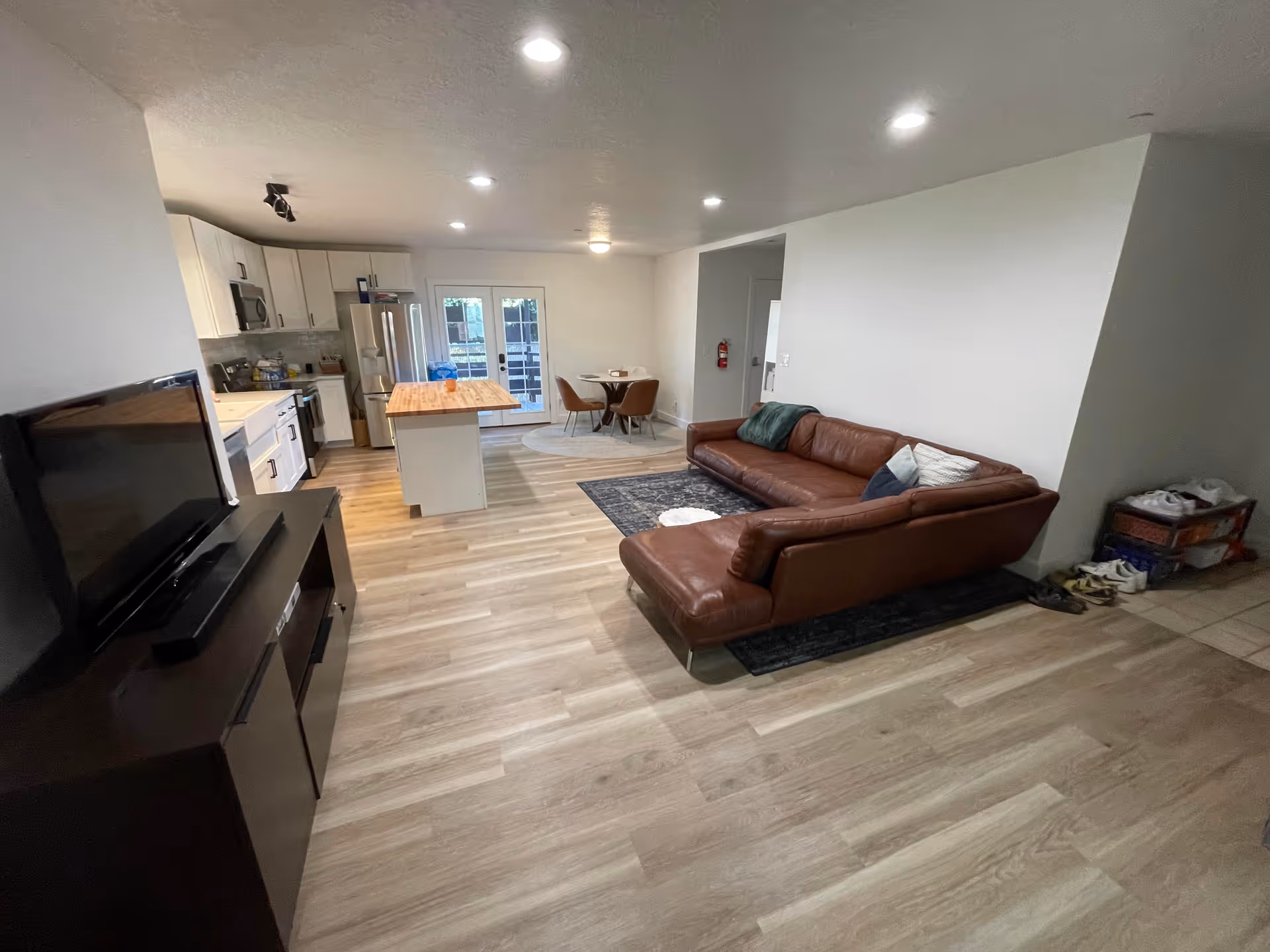 Open living space with a brown leather sectional sofa on a dark rug, a flat-screen TV on a dark TV stand, a kitchen area with white cabinets, stainless steel appliances, and a wooden island countertop, and a small round dining table with four chairs near glass double doors leading outside.