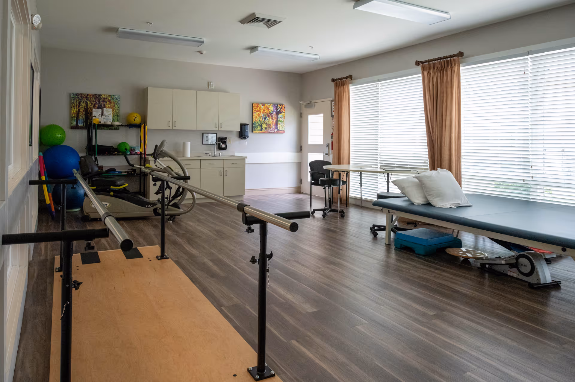 A rehabilitation or physical therapy room with parallel bars for walking exercises, an exercise bike, therapy balls, a treatment table with pillows, a small table with a chair, and cabinets along the wall. Large windows with blinds and curtains allow natural light into the room.