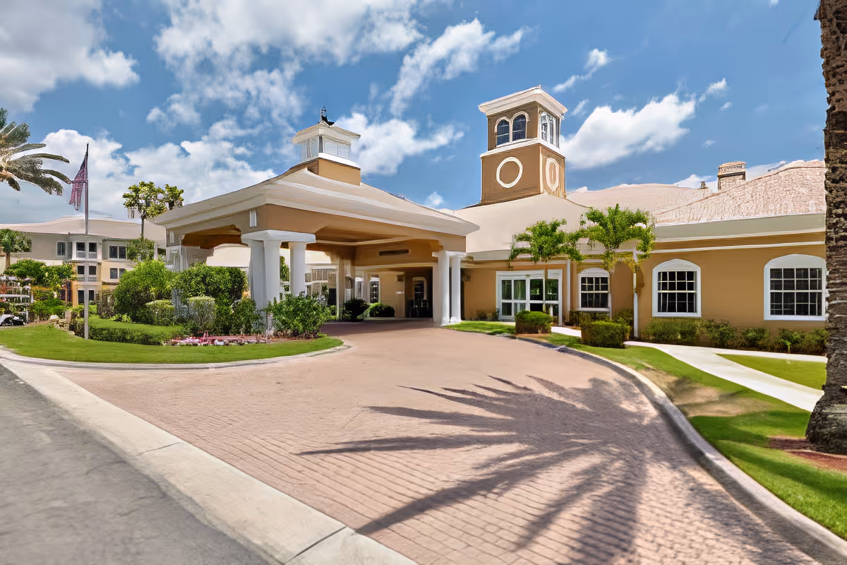 Exterior view of a senior living facility with a covered entrance, beige buildings, white columns, palm trees, and a clear blue sky with some clouds.