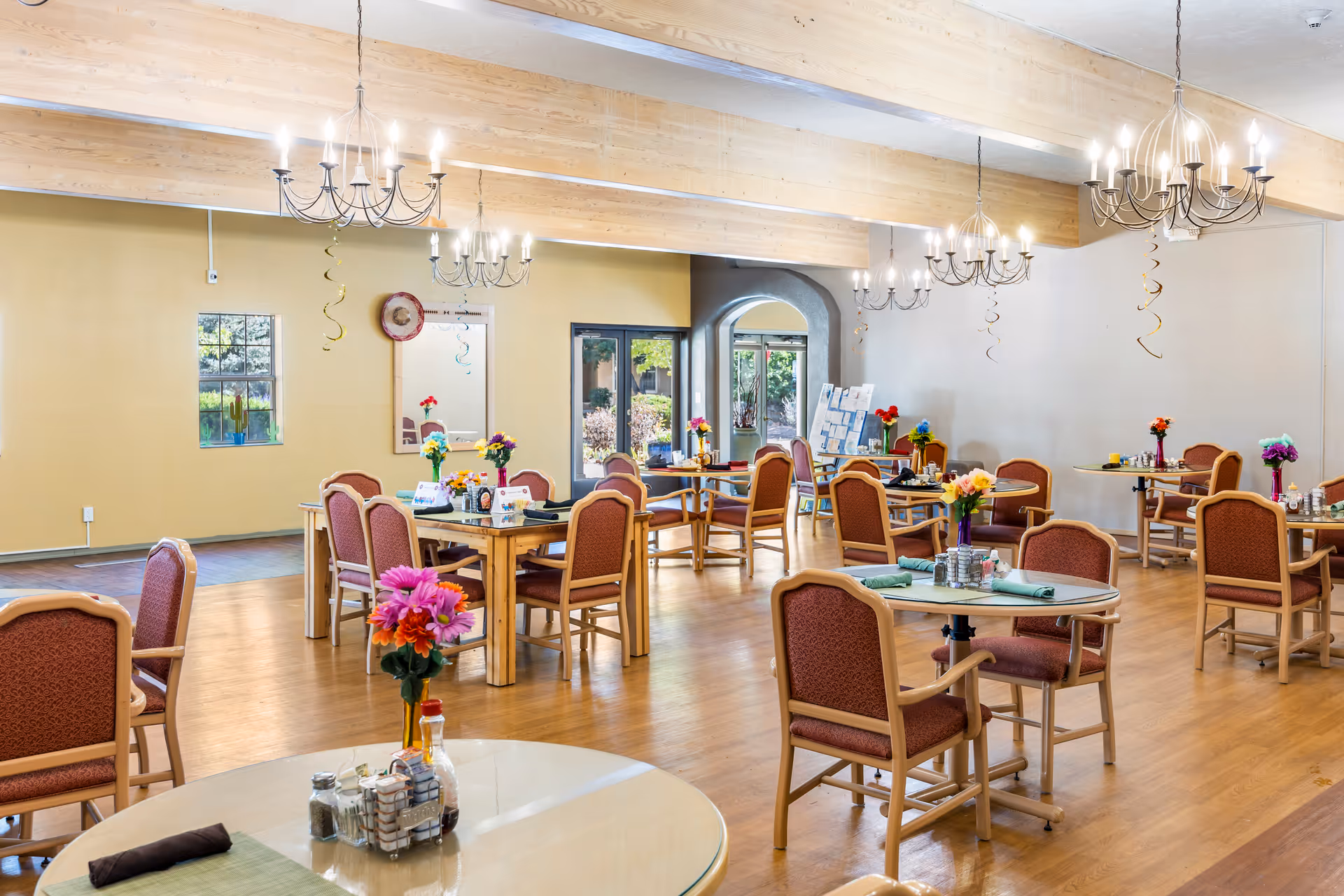 A bright and spacious dining room with several round and rectangular tables, each surrounded by wooden chairs with red cushions. The tables are decorated with colorful flower arrangements and place settings. The room features wooden beams on the ceiling, multiple chandeliers, large windows, and a glass door leading outside. The walls are painted in light colors, and the floor is wooden.