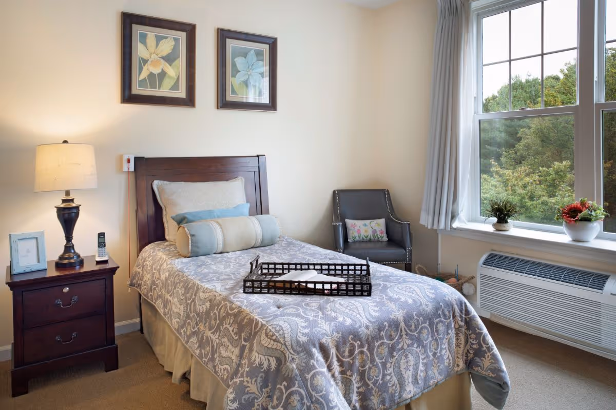 A cozy bedroom in a senior living facility with a single bed covered in a patterned bedspread and multiple pillows. A wooden nightstand with a lamp, a framed photo, and a cordless phone sits beside the bed. There is a gray armchair with a decorative pillow in the corner near a large window with white curtains, showing green trees outside. A heating/cooling unit is installed below the window, and two framed floral artworks hang on the wall above the bed.