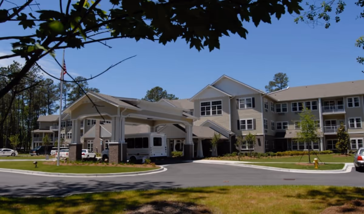 Front exterior of a three-story senior living facility with a covered porte-cochère, parked shuttle van, and an American flag.