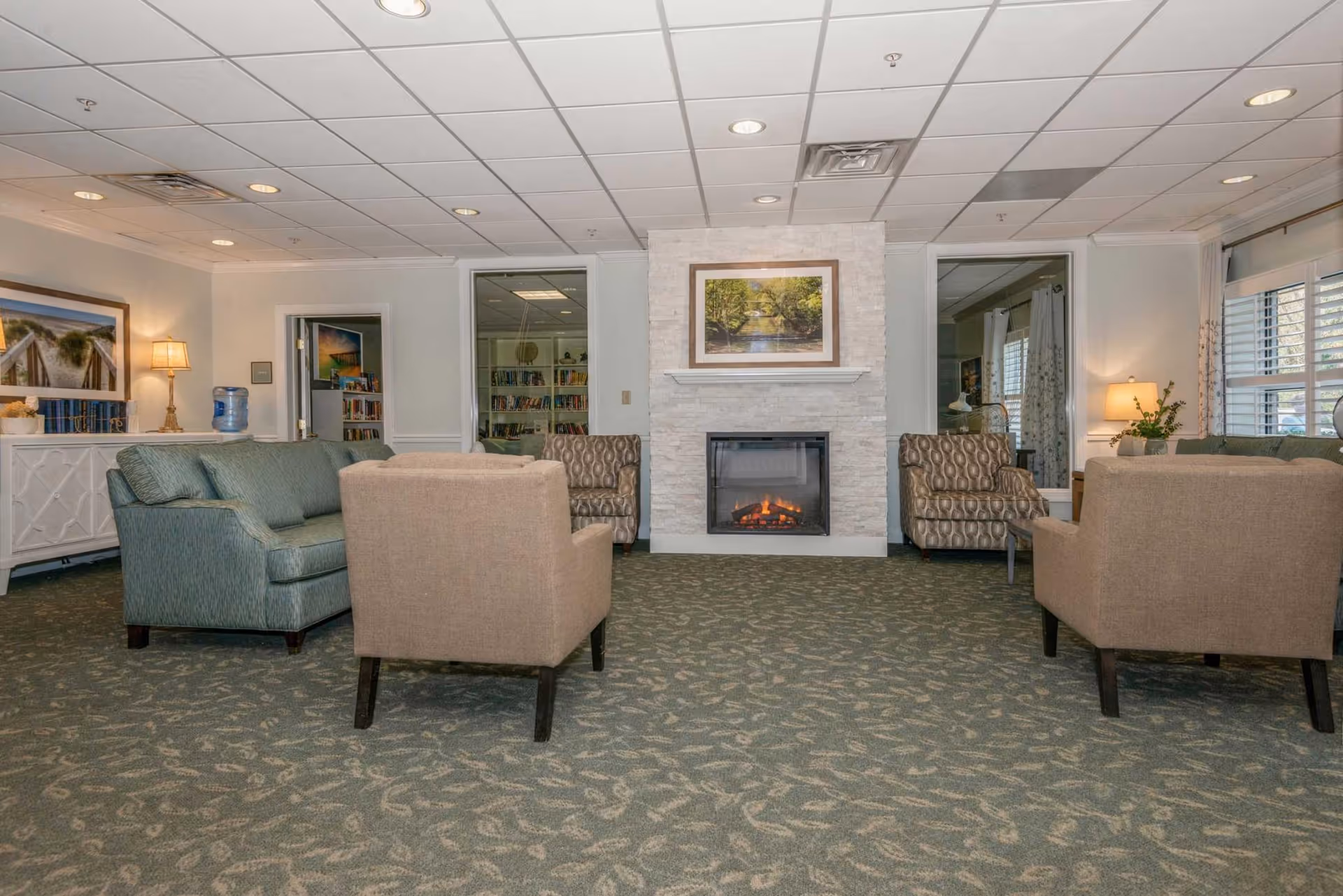 A comfortable common sitting room with sofas and armchairs arranged around a central fireplace topped by a framed landscape picture.