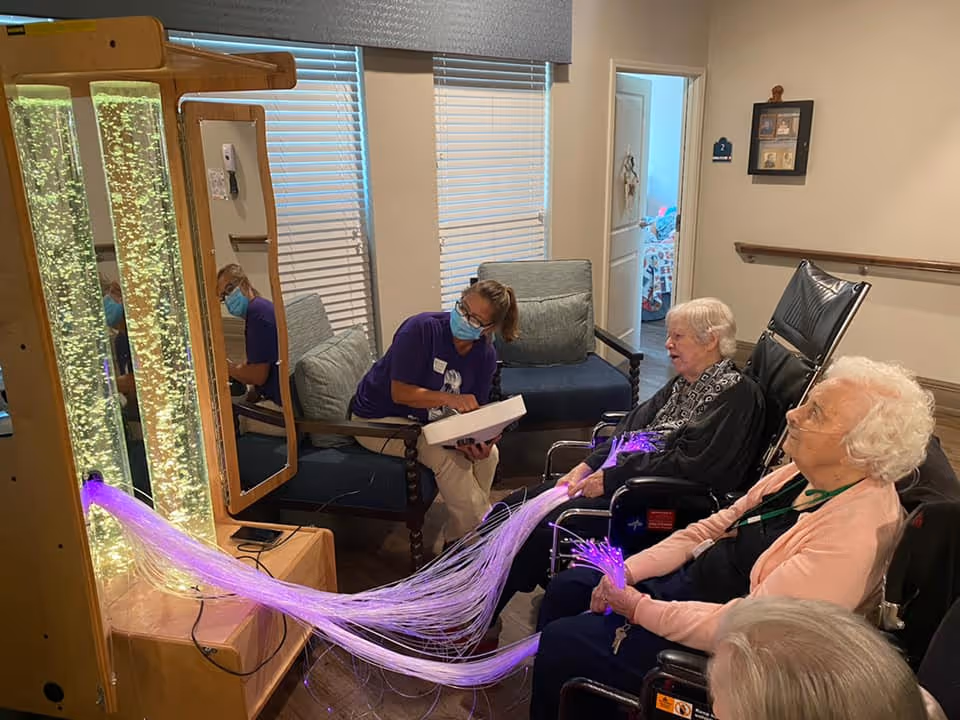 A caregiver wearing a mask interacts with three elderly women seated in wheelchairs in a cozy room. The women are holding and touching fiber optic sensory lights that emit a purple glow. Behind the caregiver, there are two windows with blinds and a cushioned chair. The room has light-colored walls and a framed picture on the wall.