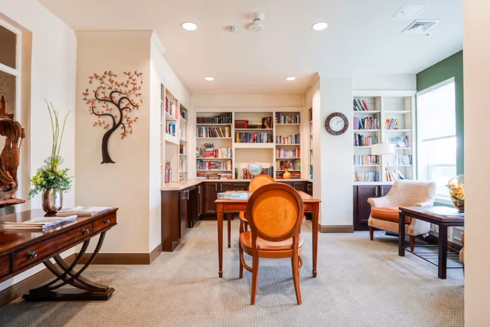 A bright and cozy reading room with built-in bookshelves filled with books, a wooden table with two chairs, a comfortable armchair near a large window, a wall clock, and decorative items including a metal tree wall art and a vase with flowers on a side table.